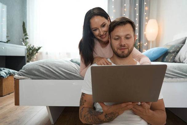 Young couple in bedroom is happy to look at photo on laptop, guy and his girlfriend are in good mood - Foto, immagini
