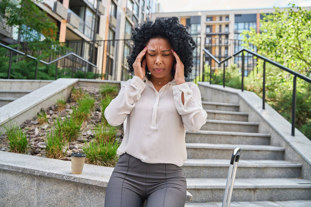 Multiracial woman in pain clutches her temples with force and winces from severe headache on yard steps - 写真・画像