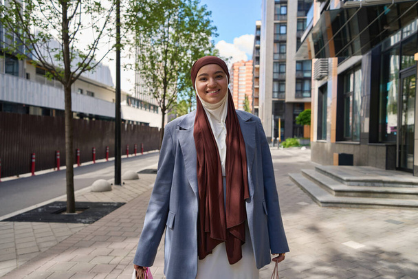 Nainen hijabissa kävelee aurinkoisella kadulla ostoksilla käsissään. - Valokuva, kuva