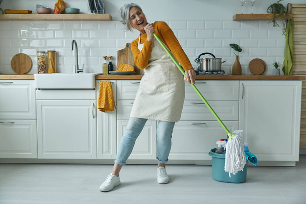 Playful senior woman carrying mop like microphone while standing at the domestic kitchen - Foto, afbeelding