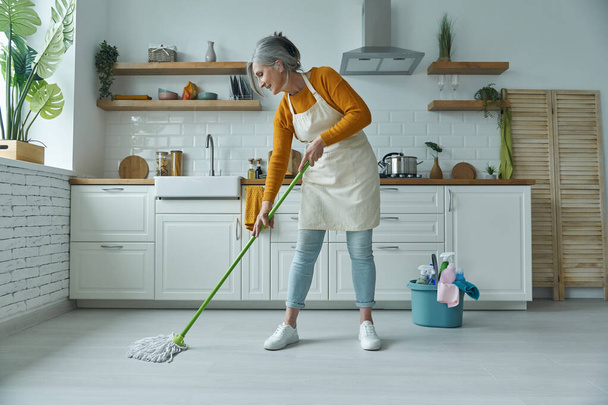 Full length of confident senior woman cleaning floor with mop while standing at the domestic kitchen - Фото, изображение