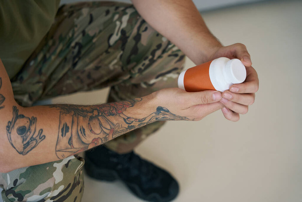 Member of military sitting at home and keeping bottle of antidepressant pills in hands - Photo, Image