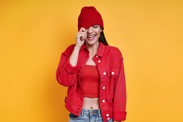 Playful young woman adjusting her hat and smiling against yellow background - Foto, Imagem
