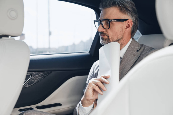 Mature businessman holding papers near face and looking thoughtful while sitting in the car - Foto, immagini