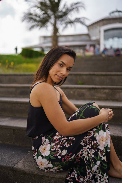 posing sitting on a bleachers a young brunette woman with short straight hair, wears a flower print skirt, summer fashion, lifestyle and natural beauty - Fotografie, Obrázek