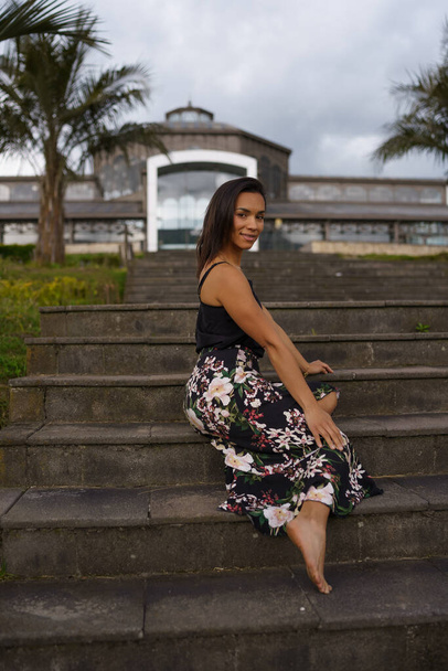 female model posing on a bleachers in the city wearing a long flower print skirt and barefoot, brunette latin woman with short black hair, lifestyle - Photo, image
