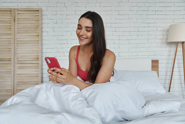 Happy young woman using smart phone while sitting in bed at home - Fotografie, Obrázek