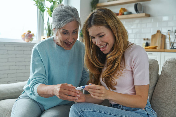 Happy woman and her senior mother looking at the pregnancy test while sitting on the couch at home - Foto, immagini