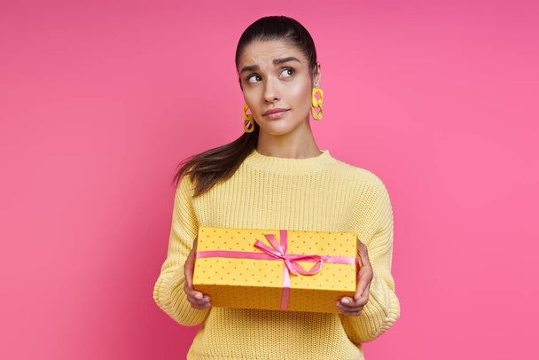 Thoughtful young woman holding gift box and looking away against colored background - Photo, Image