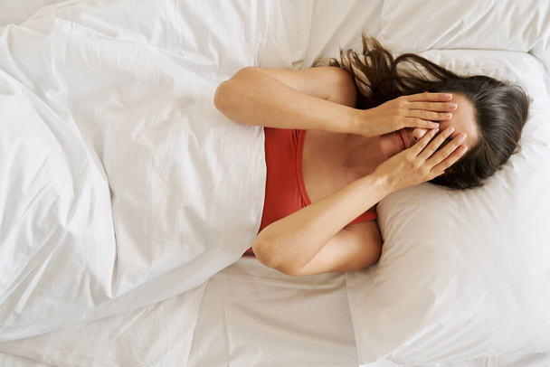 Top view of depressed young woman covering face with hands while lying in bed - Foto, Bild