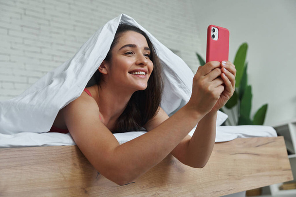 Beautiful young woman covering with blanket while using smart phone in bed at home - Fotó, kép
