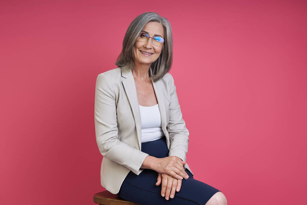 Confident senior woman in formalwear smiling while sitting against pink background - Photo, Image