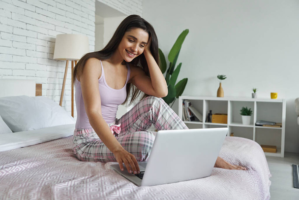 Beautiful young woman using laptop while sitting on bed at home - Foto, Bild