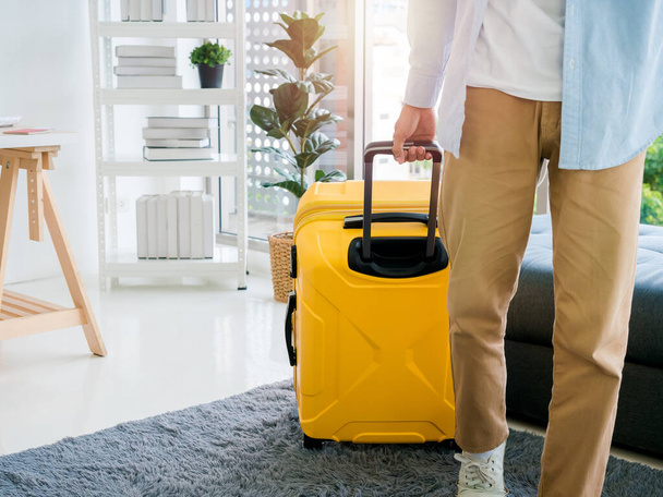 A man with yellow luggage. A male in denim shirt and brown trousers walking while holding a suitcase at the handle in office room at home. Summer holiday travel vacation concept. Time to journey. - Φωτογραφία, εικόνα