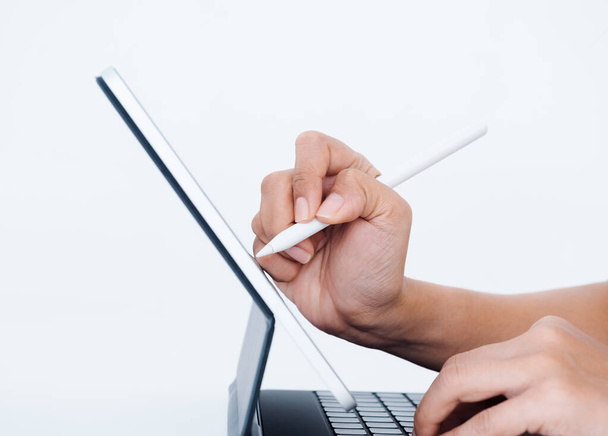Hand holding white electronic pen or pencil, writing or drawing on digital tablet screen while typing on keyboard computer isolated on white background, side view. Work with technology. - 写真・画像