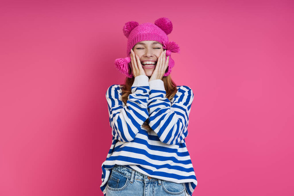 Playful young woman in pink hat touching face and smiling while standing against colored background - Fotografie, Obrázek