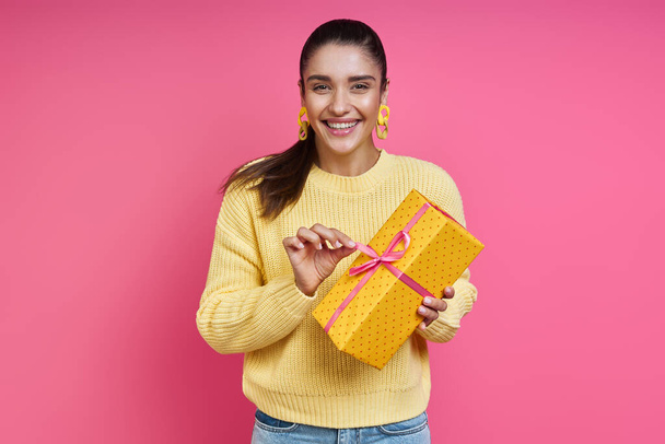 Happy young woman opening gift box while standing against colored background - Фото, зображення