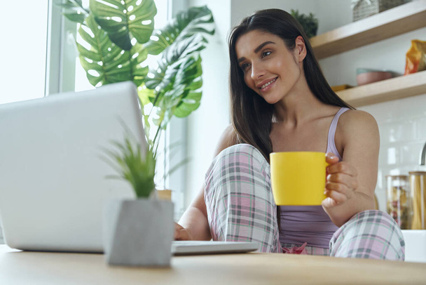 Красива молода жінка насолоджується ранковою кавою і використовує ноутбук на домашній кухні
 - Фото, зображення