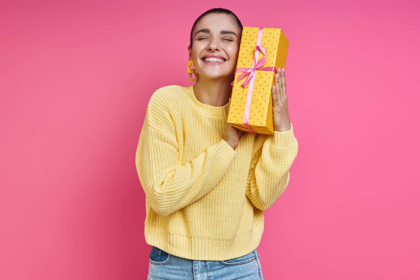 Happy young woman holding gift box near face and smiling against colored background - Foto, Imagem