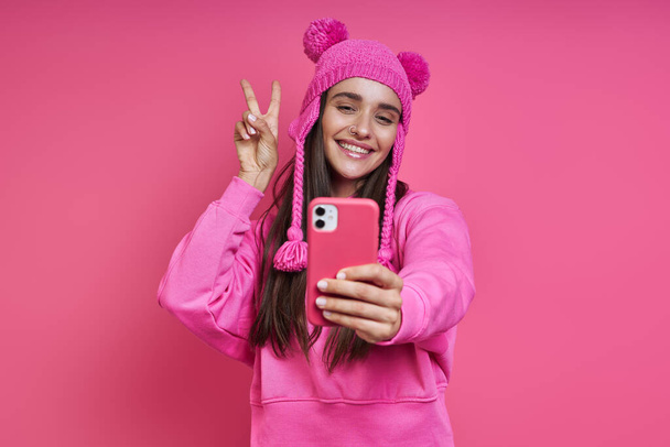 Beautiful young woman in funky hat making selfie and gesturing against pink background - Фото, изображение