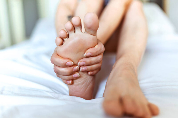 Біль у ногах жінка сидить на ліжку, тримаючи ноги вдома, болючі ноги і розтягуючи м'язи втому, щоб полегшити біль. Концепція здоров'я
. - Фото, зображення