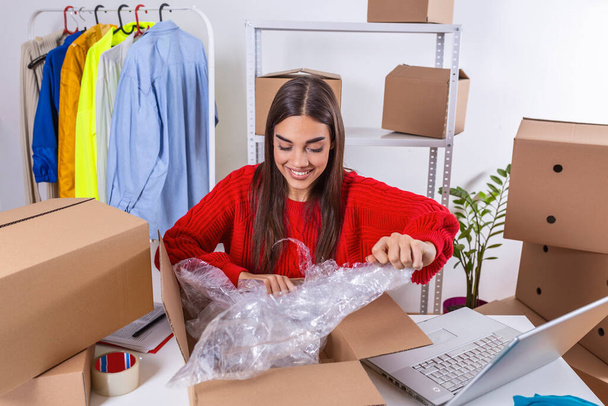 Working woman at online shop. She is wearing casual clothing and packaging goods for delivery. Owener of small business packing product in boxes, preparing it for delivery,checking on laptop address - Foto, Imagem