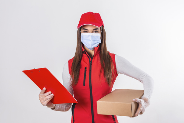 Delivery person delivering packages holding clipboard and package smiling happy in red uniform. Beautiful young woman wearing medical mask and gloves professional courier isolated on white background - Photo, Image