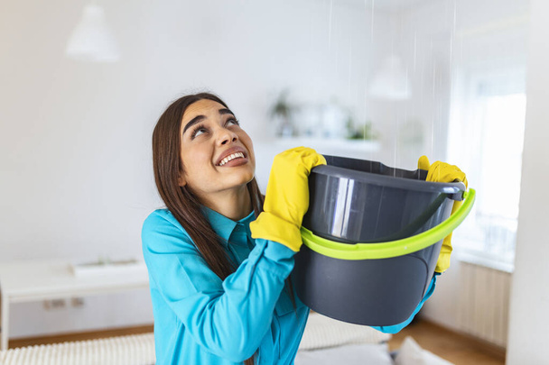 Shocked Woman Looks at the Ceiling While Collecting Water Which Leaks in the Living Room at Home. Worried Woman Holding Bucket While Water Droplets Leak From Ceiling in Living Room - Zdjęcie, obraz