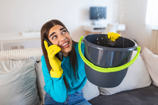 Šokovaná žena volající instalatér při shromažďování vody vytékající z strop pomocí náčiní - Fotografie, Obrázek