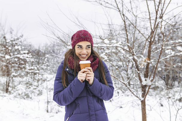 Pretty young woman in winter outfit holding disposable cup filled with hot coffee or tea. Girl holding mug of hot beverage in her hands and walks outdoors in winter. - Foto, afbeelding