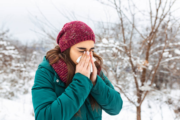 Young woman blowing in a tissue in a cold winter with a snowy mountain in the background. Portrait of a woman with tissue in hands looking away. Girl has the flu and running nose - 写真・画像