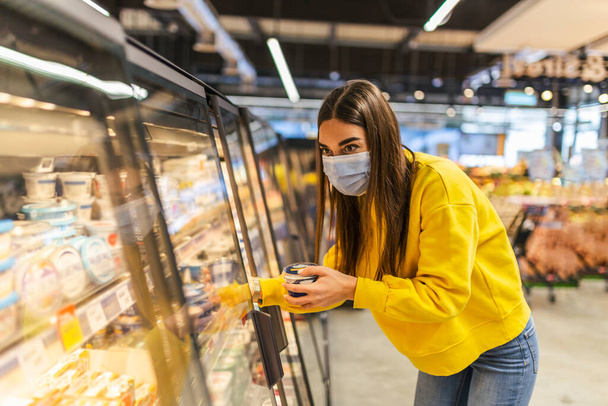 Coronavirus zatürree salgını sırasında tek kullanımlık tıbbi maske takan genç bir kadın süpermarkette alışveriş yapıyor. Salgın sırasında tedbirlerin korunması ve önlenmesi. - Fotoğraf, Görsel