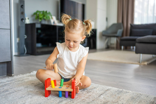 Söpö valkoihoinen pikkutyttö leikkii lattialla kotona ekopuisilla leluilla. Montessorin lelu. Lapsi pelaa opettavaisia pelejä. Laadukas kuva - Valokuva, kuva