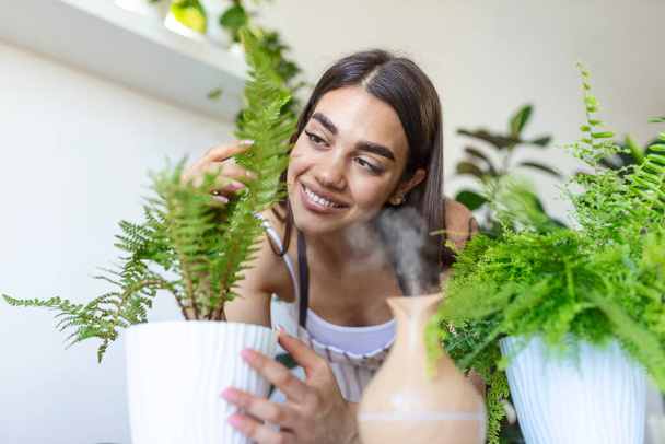 Woman caring for plants next to steam aroma oil diffuser on the table at home, steam from humidifier. Humidification of air in apartment during the period of self-isolation due to coronavirus pandemic - Photo, Image