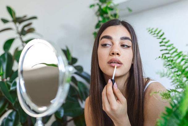 Beautiful young woman applying lip gloss with applicator in front of a mirror at home - Photo, image