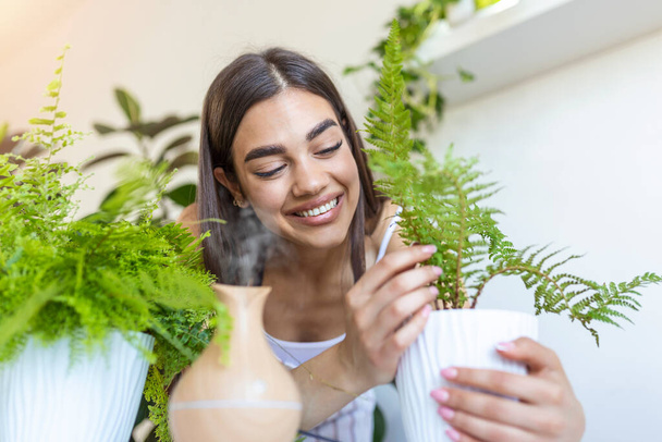 Woman caring for plants next to steam aroma oil diffuser on the table at home, steam from humidifier. Humidification of air in apartment during the period of self-isolation due to coronavirus pandemic - Φωτογραφία, εικόνα