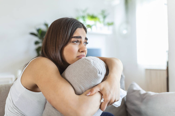 Young woman hugging pillow with depression and crying on sofa at home. Lonely millennial lady feeling stressed and hopeless, suffering from mood disorder or having psychological problem - 写真・画像