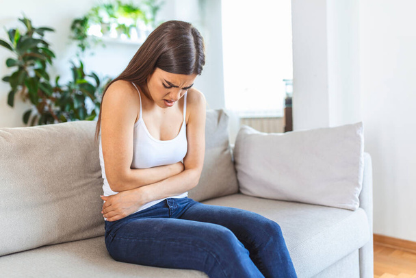 Woman in painful expression holding hands against belly suffering menstrual period pain, lying sad on home bed, having tummy cramp in female health concept - Foto, Imagen