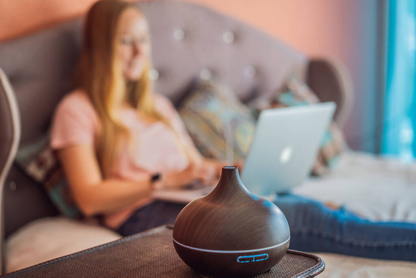 A woman sits on a bed and works on a laptop. Aroma diffuser next to the bed. - 写真・画像