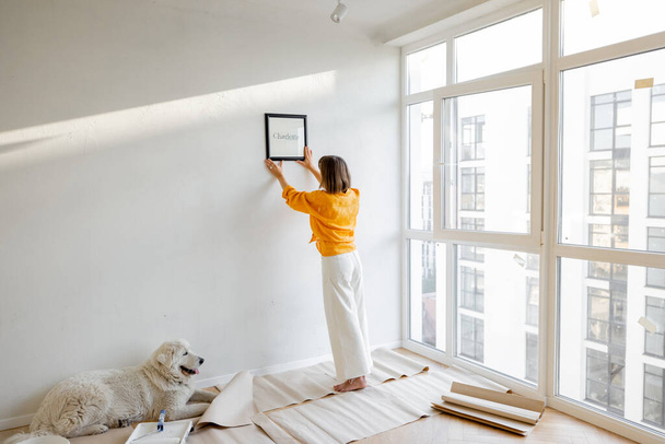 Young woman hanging picture frame in room, decorating her newly renovated apartment, stands with her dog in white room - Photo, Image