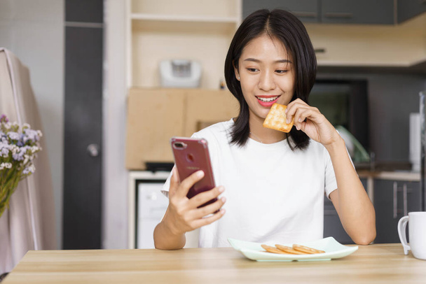 Концепция домашнего отдыха, молодая женщина ест крекеры и серфинг социальных сетей на смартфоне дома. - Фото, изображение