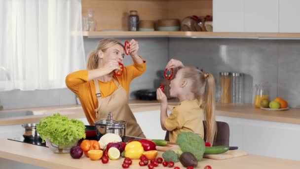 Funny culinary. Playful carefree mother and her little daughter fooling around, playing with fresh pepper slices at kitchen, grimacing and laughing, tracking shot, slow motion, free space - Video