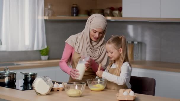Een beetje hulp. Schattig meisje helpen haar moslim moeder in hijab om ontbijt te koken, kloppen eieren met melk in kom en glimlachen, vrije ruimte - Video