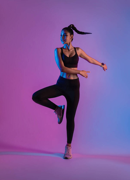 Atleta asiática deportista saltando danza como parte de la quema de grasa entrenamiento en el estudio de fitness fondo de neón. Mujer haciendo ejercicio con cardio en el gimnasio. - Foto, imagen