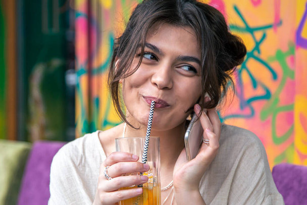 Funny young woman talking on the phone with a glass of lemonade on a colorful background. - Фото, изображение