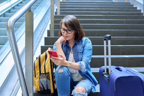 Woman passenger with suitcase backpack sitting on steps in modern building. Female with smartphone luggage at train station, airport. Passenger transport, transportation, trip, journey, travel, people - Photo, image