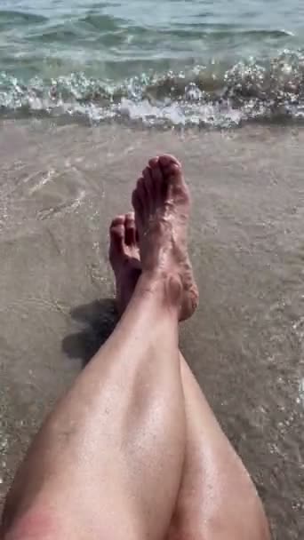 first person view of the crossed legs of a man sitting on the seashore on a white sandy beach, with the waves crashing over them, resting on the seashore on holiday, vertical - Filmagem, Vídeo