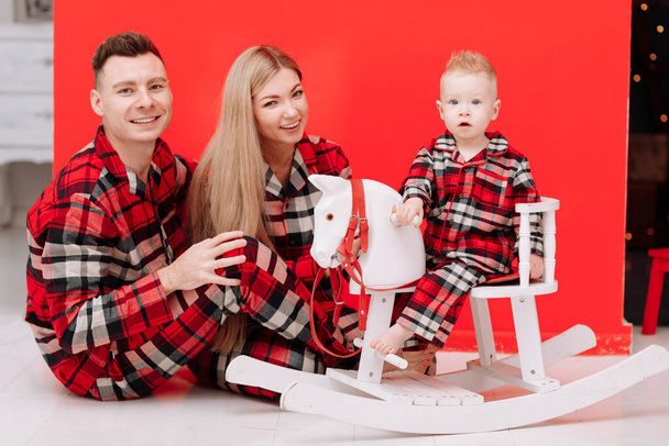 smiling young mom, dad are playing with baby boy on wooden rocking horse on the red background. happy family with little child in red checked pyjamas having fun together. Childcare, childhood - 写真・画像