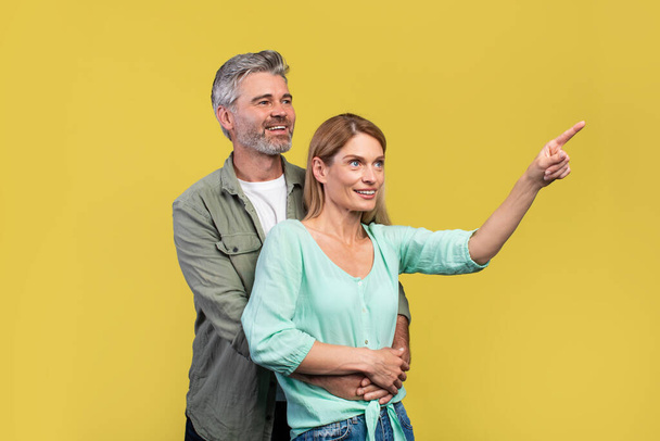 Check this cool offer. Positive mature spouses embracing and pointing aside at free space, showing empty free place for advertisement over yellow background - Foto, imagen