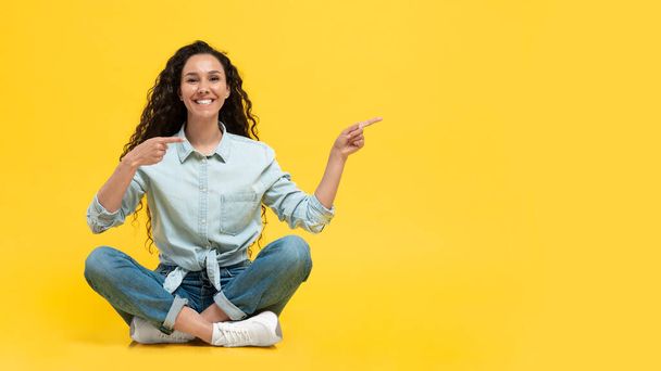 Happy Middle Eastern Woman Pointing Fingers Aside Showing Free Space Smiling To Camera Sitting In Studio Over Yellow Background. Female Advertising Yout Text. Look There. Panorama - Foto, Bild
